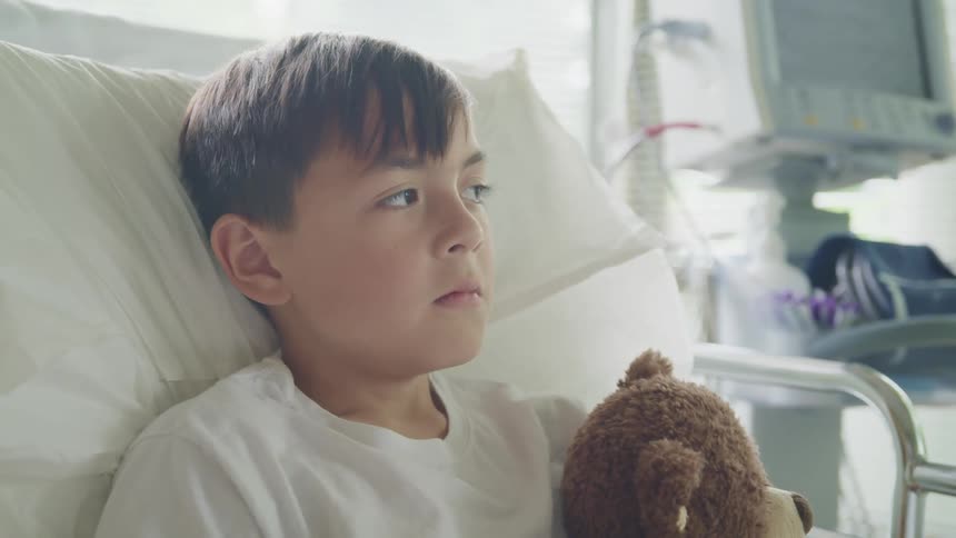 A young boy sits in a hospital bed holding a teddy bear