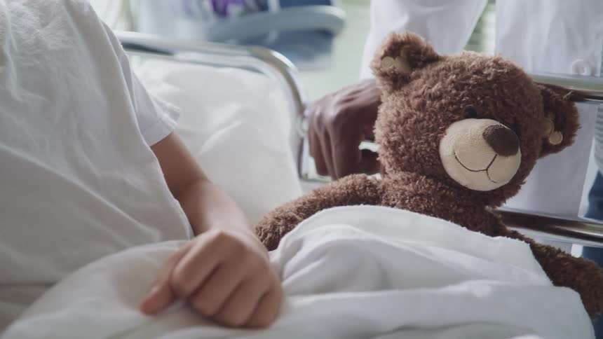 A child rests in a hospital bed next to a teddy bear as a doctor stands at bedside