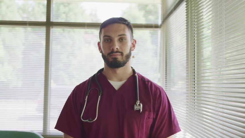 Man in maroon scrubs with stethoscope stands in front of windows in office