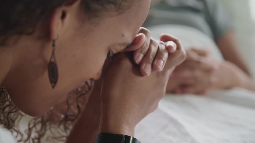 A woman prays with her head resting on her hands at the bedside of a young man in a hospital