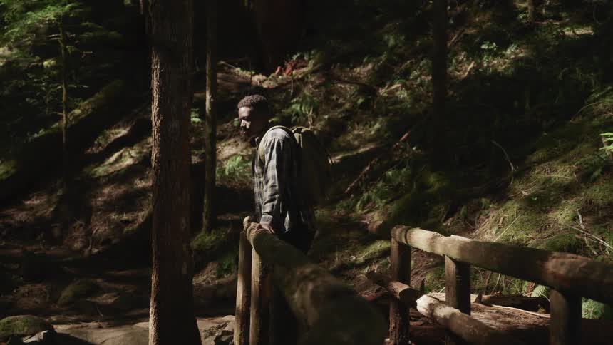 Black man leans against a wooden railing of a walkway near a hill