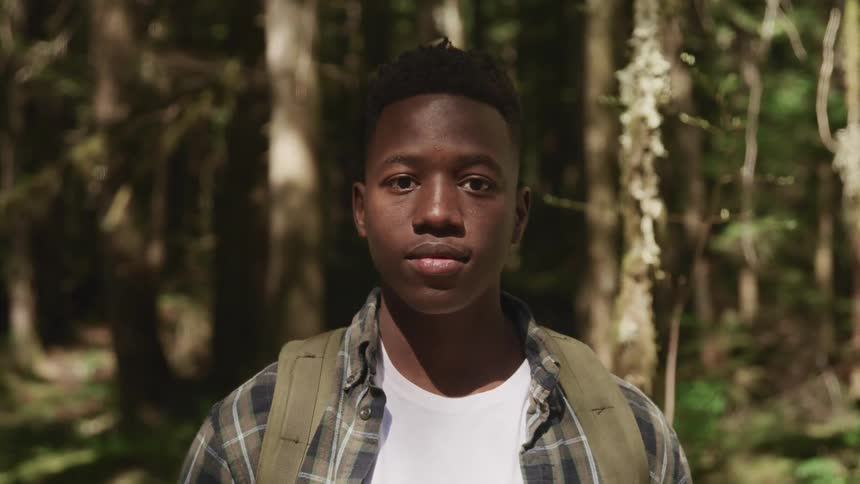 Black man with a shirt and backpack faces the camera with eye contact in the forest