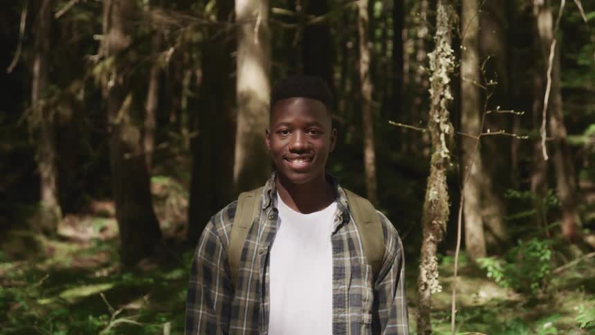 Black teenage boy with a shirt and backpack smiles broadly and faces the camera with eye contact in the forest