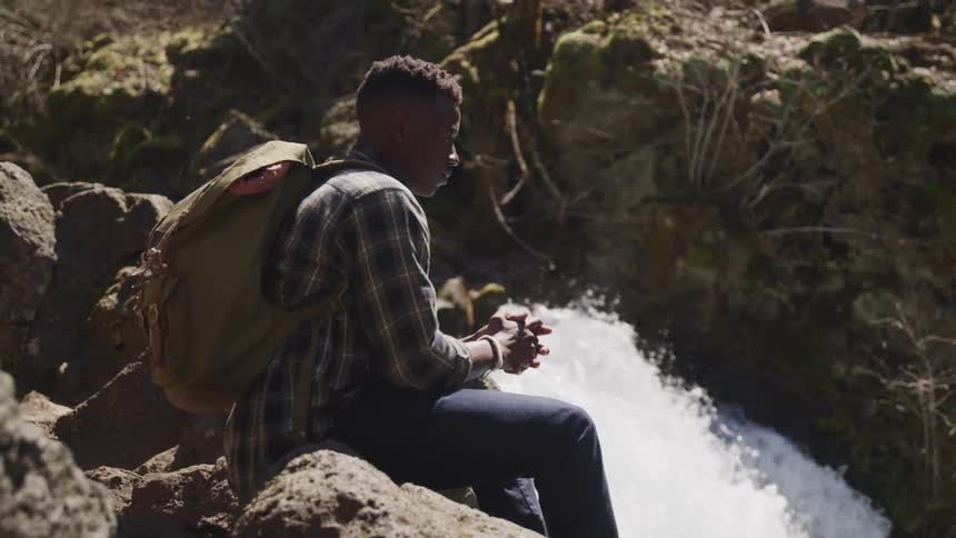 Young man wearing a backpack sits close to a waterfall