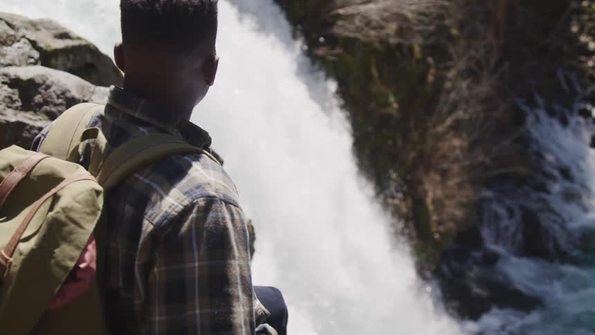 Black man with a shirt and a brown backpack watches a waterfall dropping from a higher to a lower point