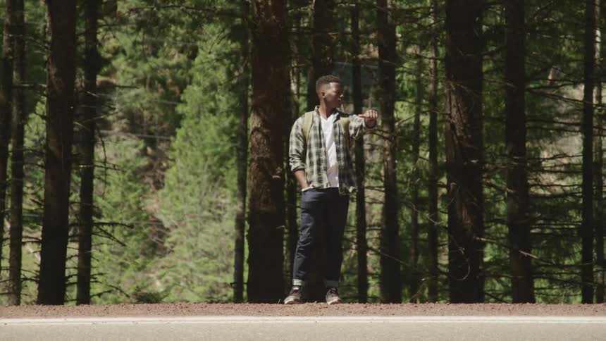Young man hitching a ride on a road next to the forest at day