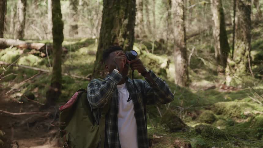 Young man wearing a backpack takes pictures of the forest with a camera at day