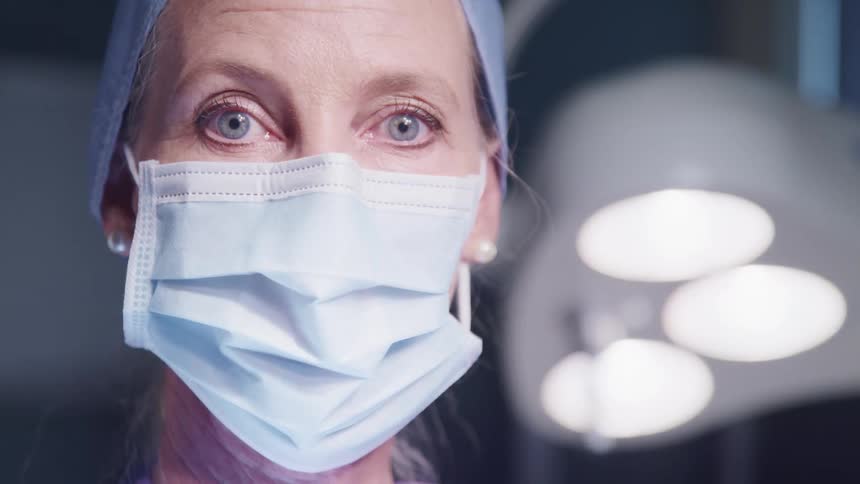 A surgeon poses in an operating room while wearing a blue surgical mask