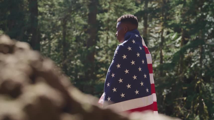 A man poses in a forest with an american flag draped over his shoulders