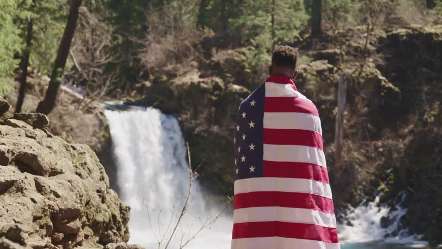 A man wearing an american flag as a cape looks at a waterfall