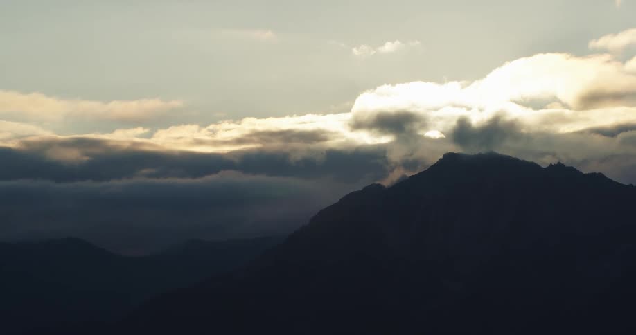 Sunlight filters through the clouds above the peaks of a mountain range at sunset