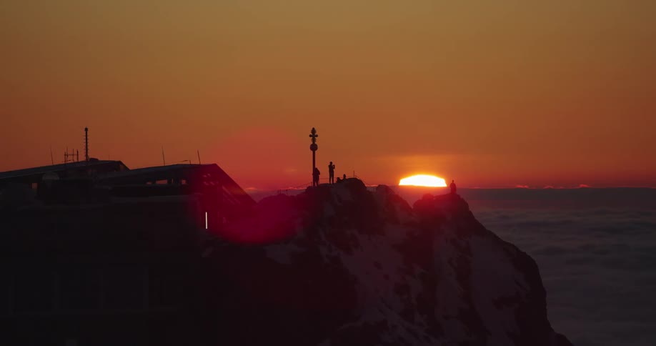 Aerial view of people standing atop of a snow covered mountain peak over the clouds at sunset
