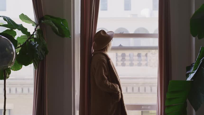 Blonde woman with a brown hat and and overcoat looks out the window