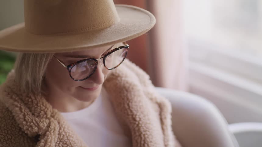 Woman with glasses a brown hat and an overcoat sits on a sofa and looks down in a room
