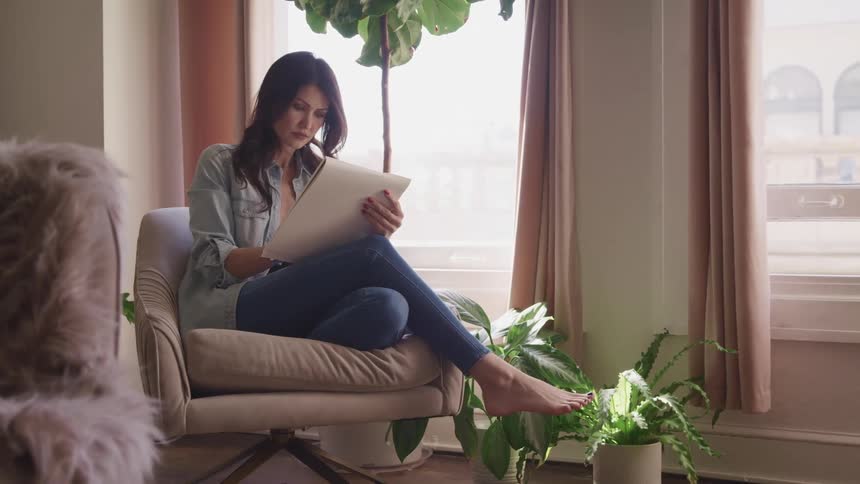 A woman sits cross legged in a pink chair by a window as she draws in her apartment