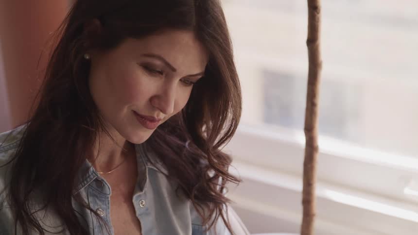 Woman with brown wavy hair and a blue shirt looks down and smiles slightly by the window in a room