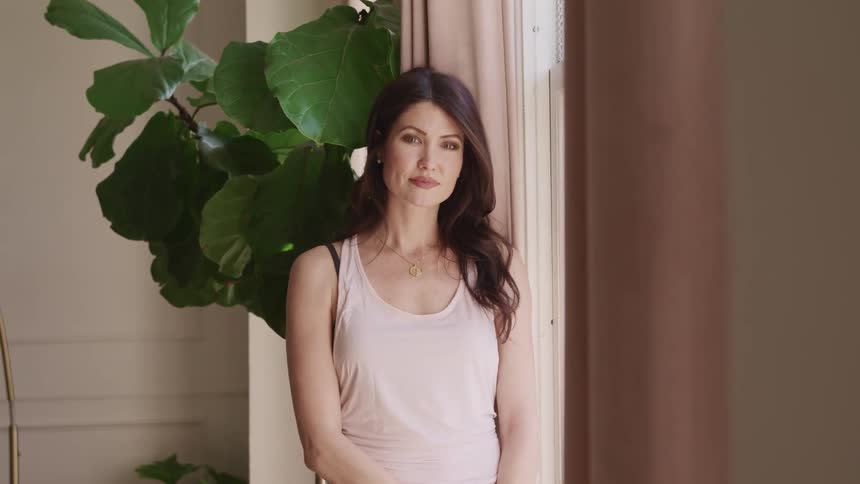 Woman standing in a room next to a window turns her face and looks at the camera smiling