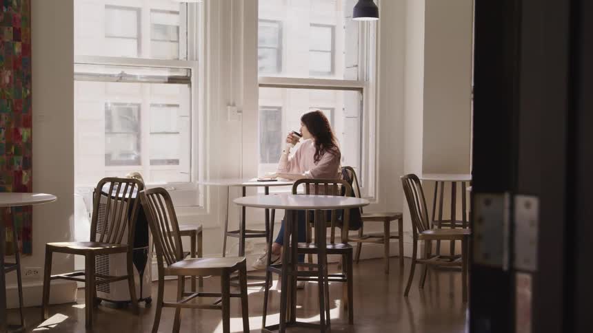 A woman sits in an empty cafe and sips on a drink sitting by the window