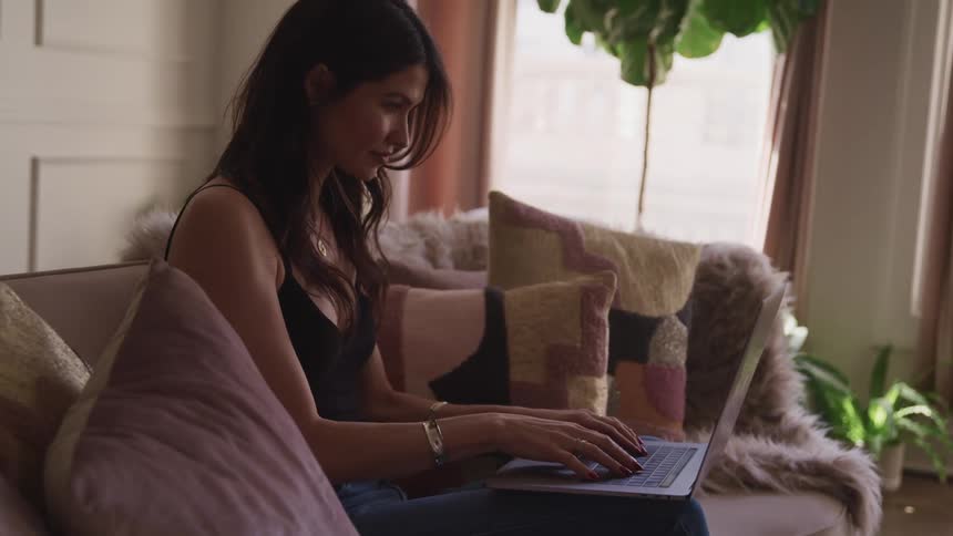 Woman with black wavy hair sits on a sofa uses a laptop in the living room