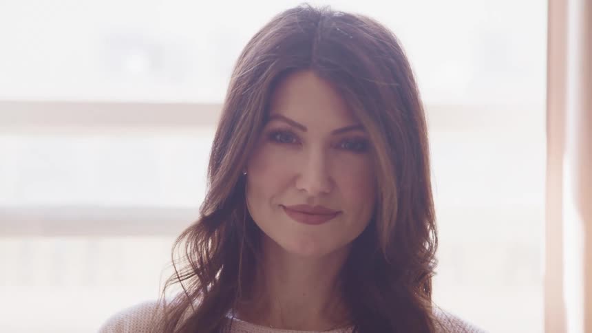 Woman with wavy brown hair and a pink sweater raises her head to look at the camera and smile slightly