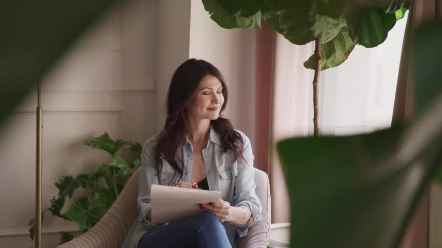 Woman sitting in a sofa on a living room writes notes with paper and pencil while looking out the window
