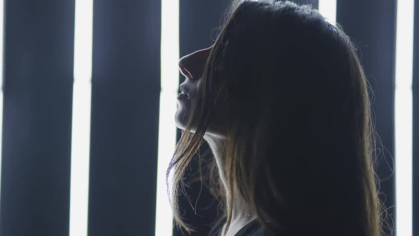 A woman with long brown hair poses in profile and raises her head up as she stands against a lighted wall