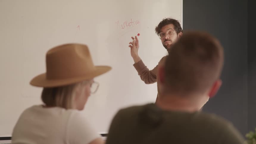 A man with a turtleneck and a marker standing in front of a whiteboard teaching