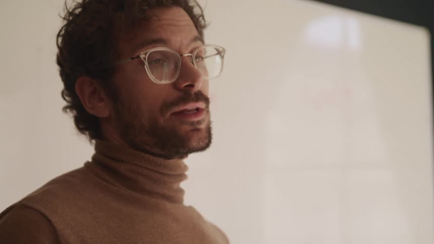 Man with a beard and clear glasses wearing a turtleneck sweater gives a lecture in front of a whiteboard