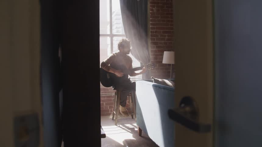 Man with a beard and glasses sits on a stool and plays a guitar in the sunlight next to the window in the living room