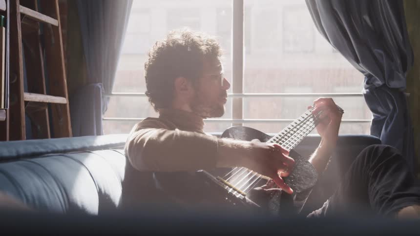 Man with a beard glasses and a brown turtleneck sweater plays a guitar as the sun shines on him