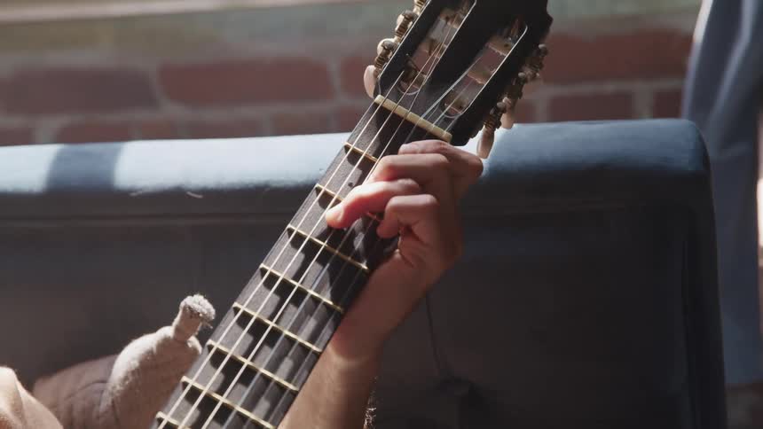 A person plays a guitar while sitting on a gray sofa