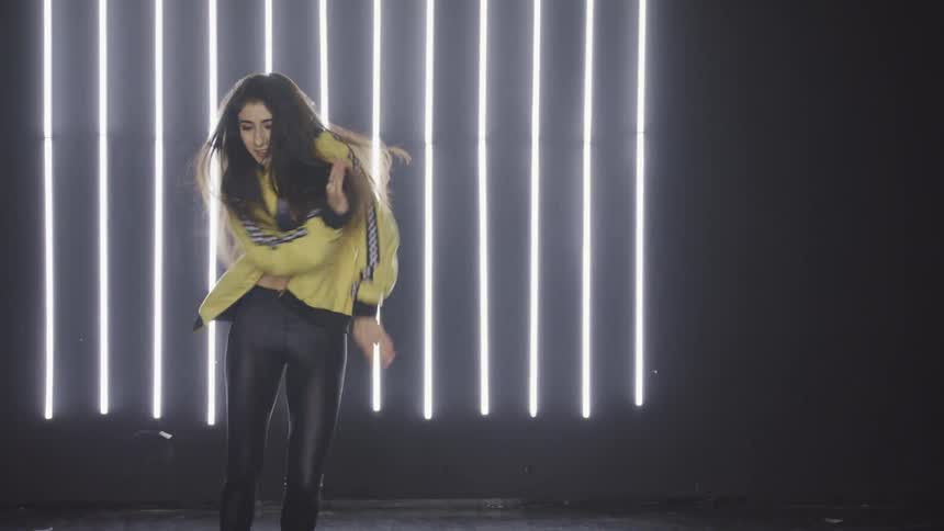 Female dancer wearing a bright yellow jacket performs dance moves on a stage in front of fluorescent lights