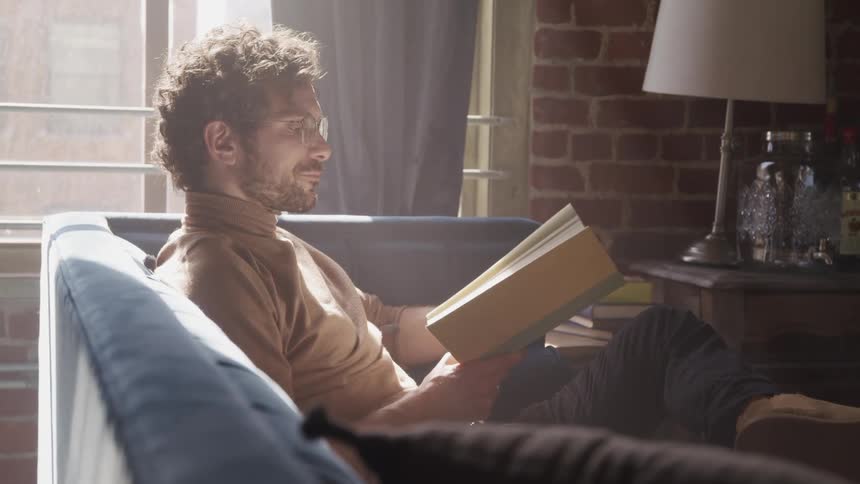 Man with a brown turtleneck sweater a beard and glasses sits on a blue sofa and reads a book