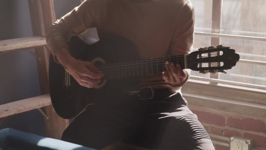 Man with a turtleneck sweater sitting on a stool plays a guitar in a room