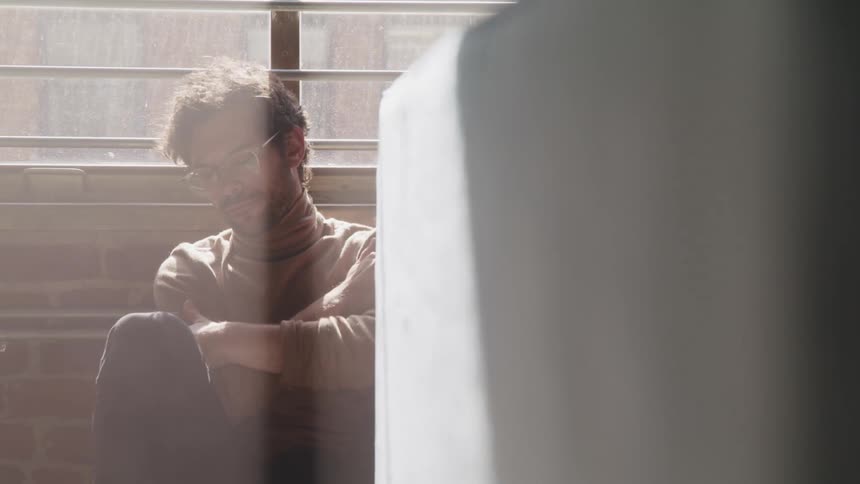 Man folds his arms as he sits on the floor in front of a window