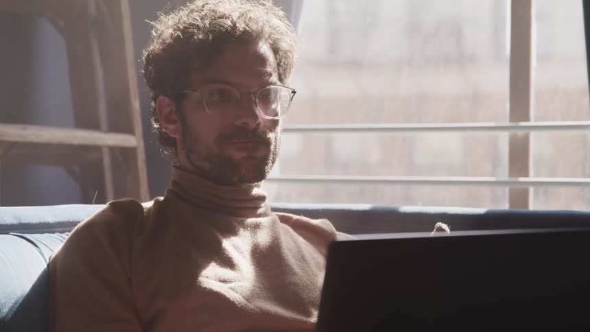 Man with a beard glasses and a brown turtleneck sweater sits on a sofa and uses a laptop in a room