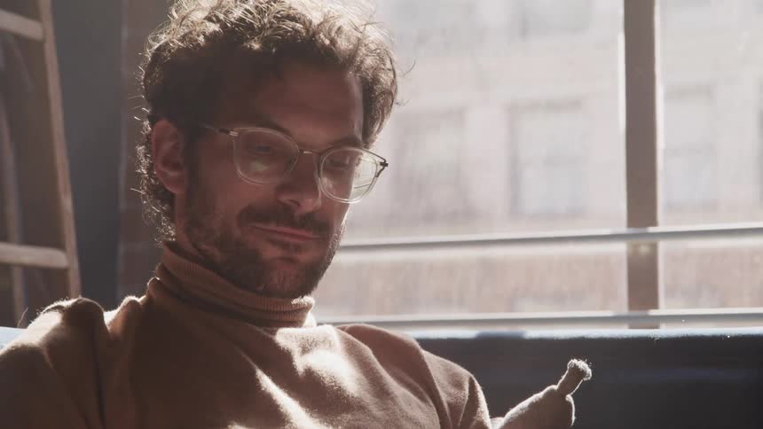 Bearded man wearing glasses sits on a room reading near to a window