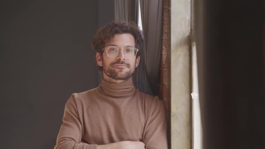 A man with a turtleneck and glasses standing by a window
