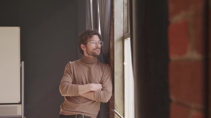 Man with a brown turtleneck sweater a beard and glasses looks out the window