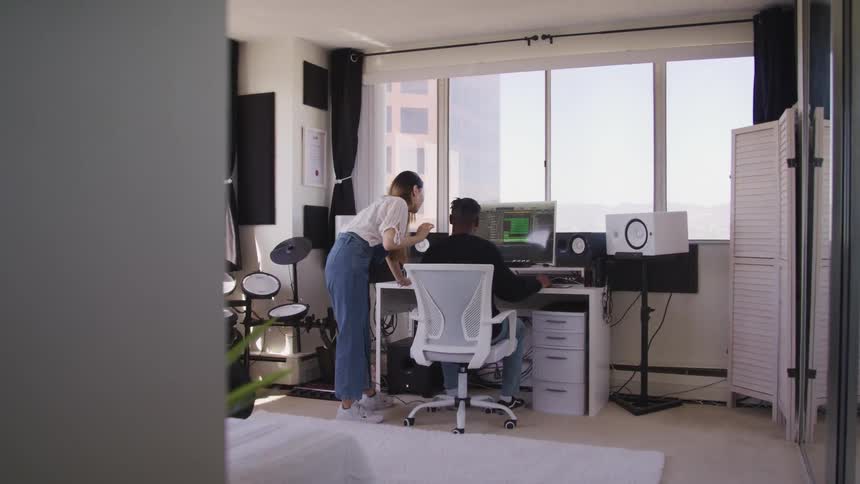 A teenager sits at a home computer desk with a woman standing beside him and leaving in towards the large computer monitor