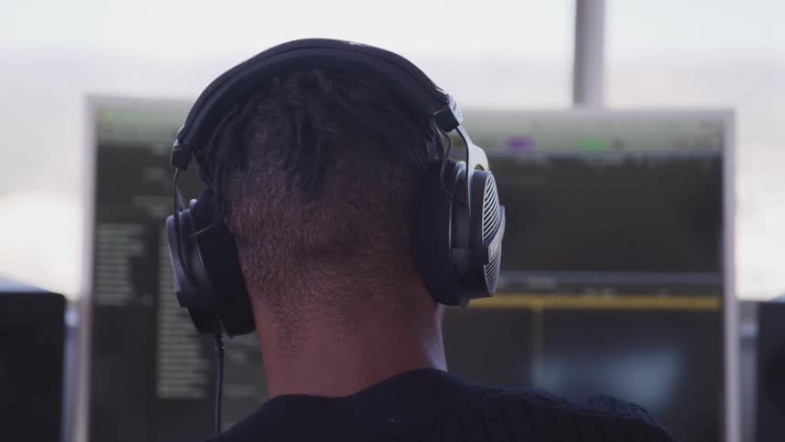 A person with headphones in front of a computer screen and speakers with a window in the background