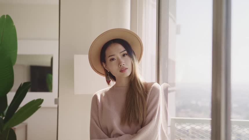 Asian woman with a brown hat and a brown sweater looks out the window and turns her gaze to the camera