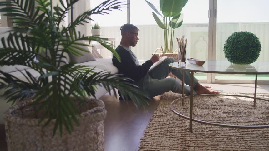 Male leaning against sofa in living room while sitting on the hardwood floor uses a pencil to draw on a sketch pad