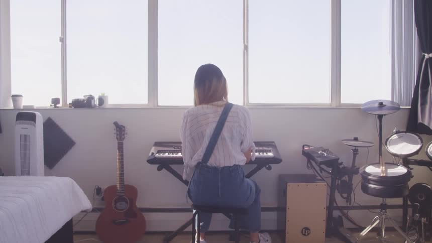 A young woman sits at a small synthesizer in a room filled with several other instruments