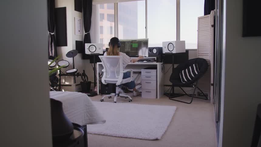 Woman sits in front of large computer screen wearing headphones surrounded by drum set and speakers