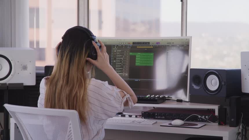 A woman in her office adjusts her headphones and looks at her computer