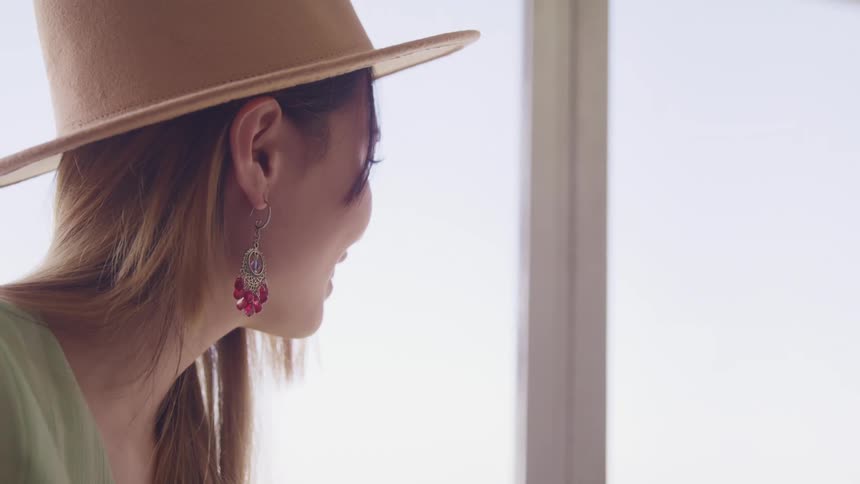 Side view of an asian woman with brown hat and earrings smiles and looking out the windows