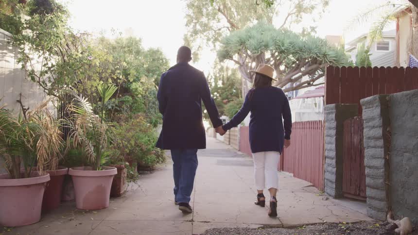 Couple walks along alley between house passing palms in large pots and fences