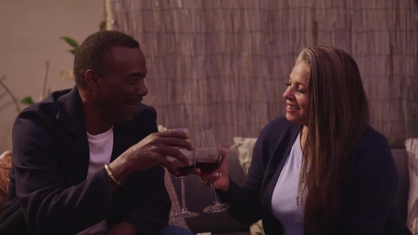 Couple smile at each other while drinking wine sitting in a couch at home