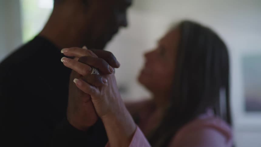 Black man with a black sweater and white woman with a pink sweater hold hands and dance romantically